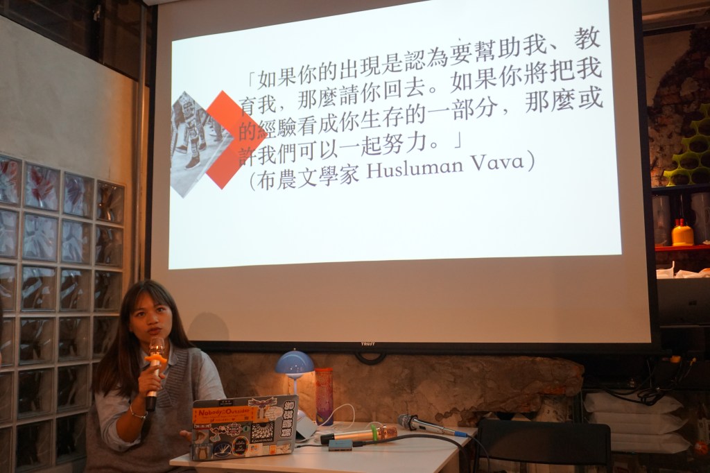 一位女性在會議室內講述,背景顯示投影畫面,文字內容與文化相關,桌上擺放麥克風和其他物品。