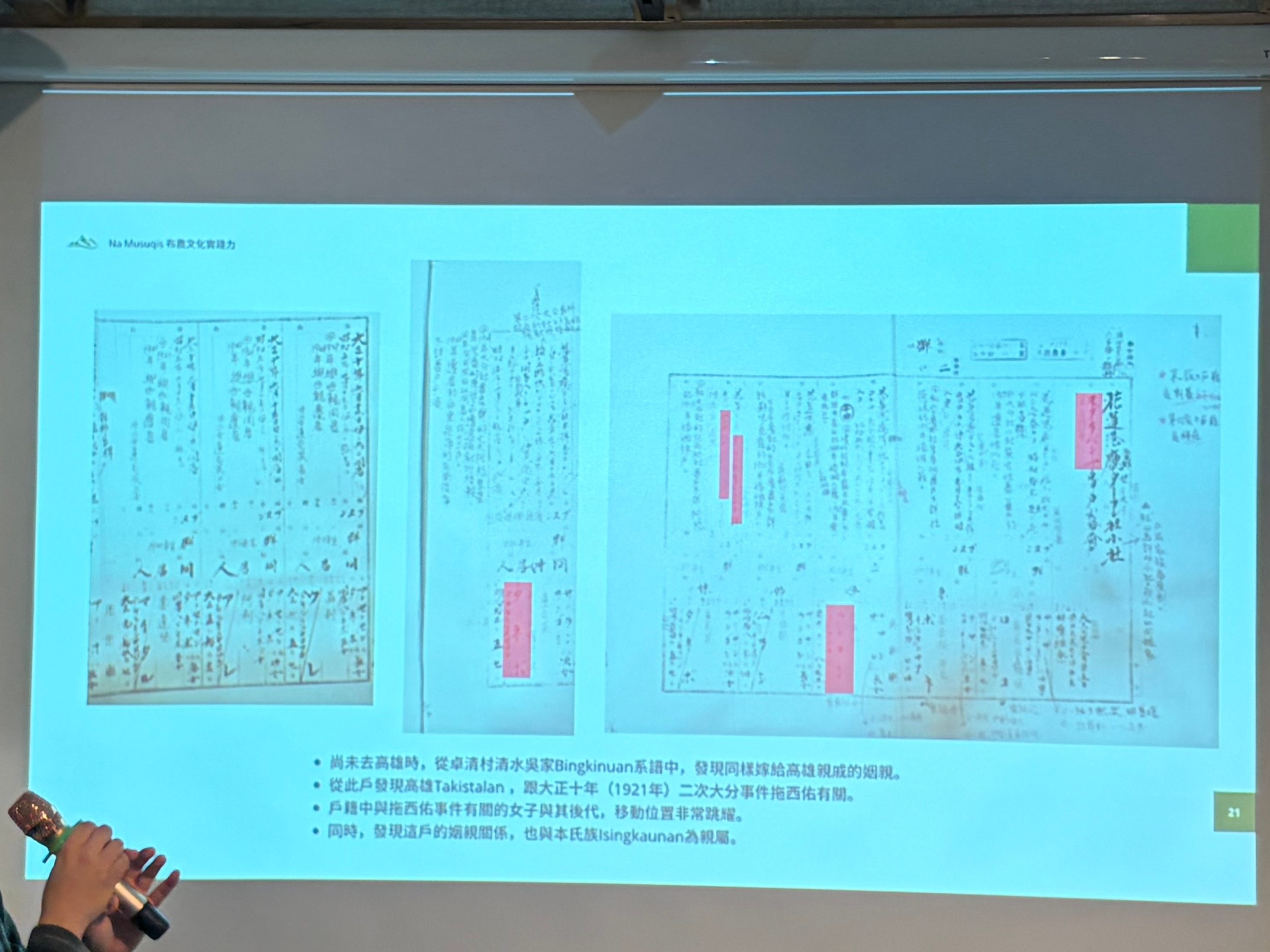 一位講者在會議上發表演說,背景是一個投影螢幕,上面展示兩張帶有標註的文件。