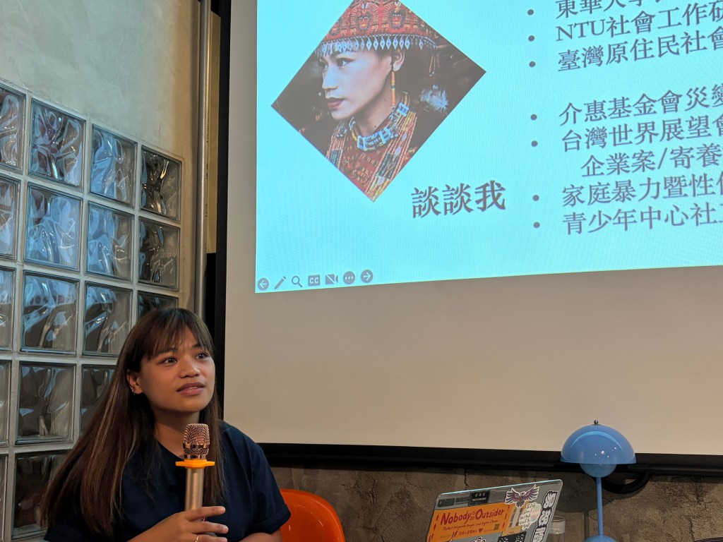 一名女性正與觀眾交談，背景為投影的簡報內容，包含台灣原住民族相關主題。