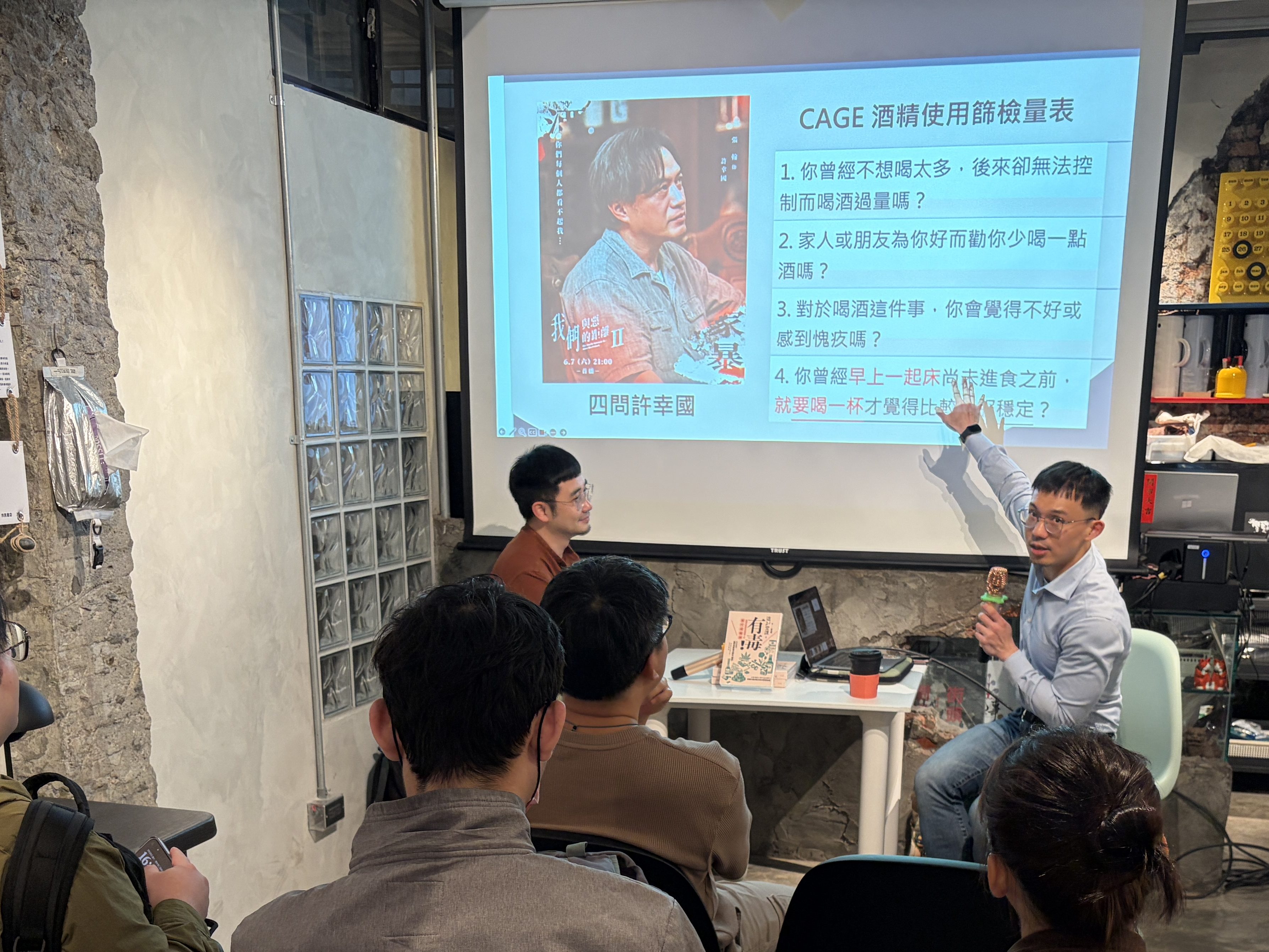 A workshop scene showing a presenter speaking to a small audience in a modern, casual setting. A projected slide displays information about the CAGE alcohol consumption assessment tool. Attendees are seated, listening attentively.