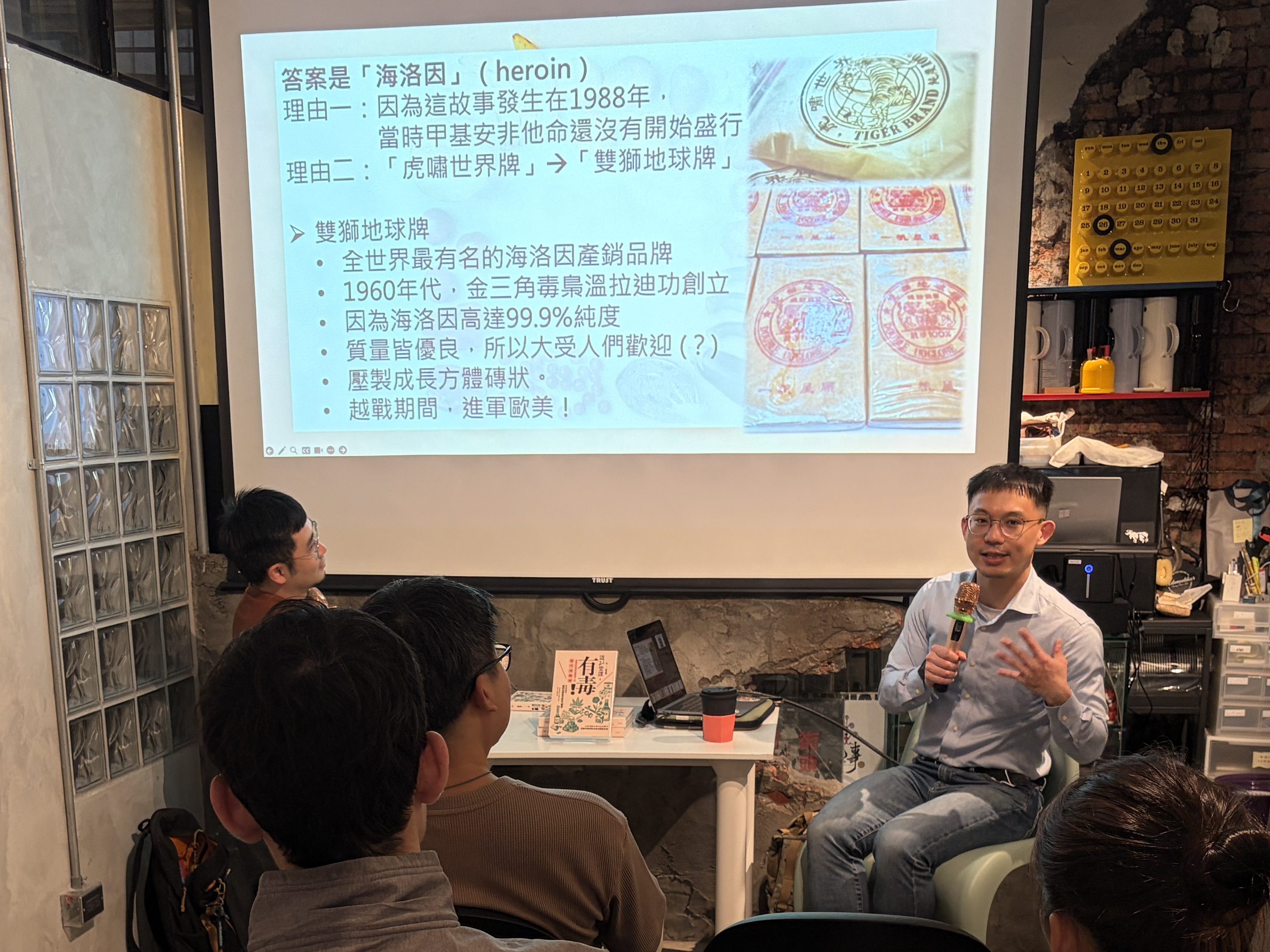 A speaker presenting to an audience in a casual setting with a projector screen displaying information. The audience, seated and engaged, includes several individuals observing the presentation.
