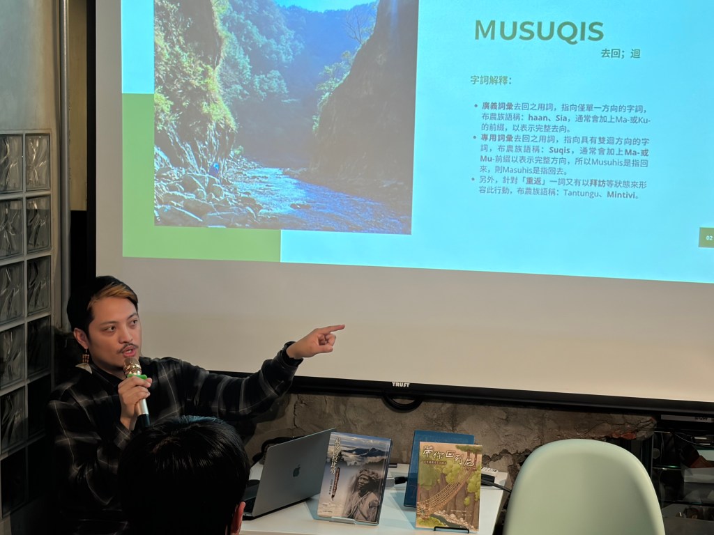 一名男性講者正在進行演講，講台上有一本書籍和一台筆記型電腦，背景是一張顯示自然風景的幻燈片，文字內容包括對字詞的解釋。