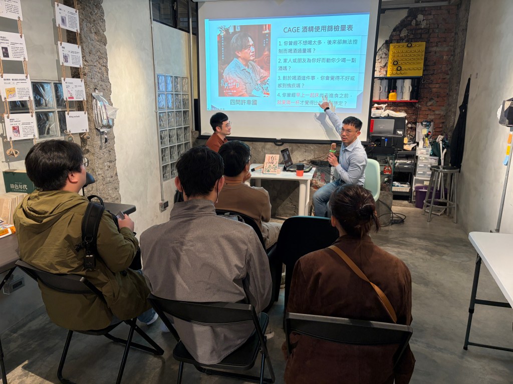 A presentation event in a modern indoor space, focusing on topics related to mental health and substance use. A speaker gestures towards a slide on the screen while an audience of five people listens attentively. The setting features exposed brick walls and contemporary furniture.