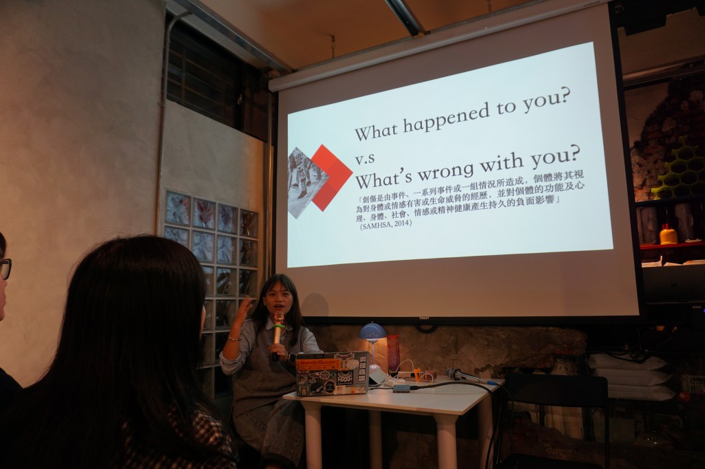 A speaker discusses a topic during a presentation, with a projector screen displaying text comparing two questions: 'What happened to you?' versus 'What's wrong with you?'. The setting is a cozy indoor space with an audience visible in the foreground.