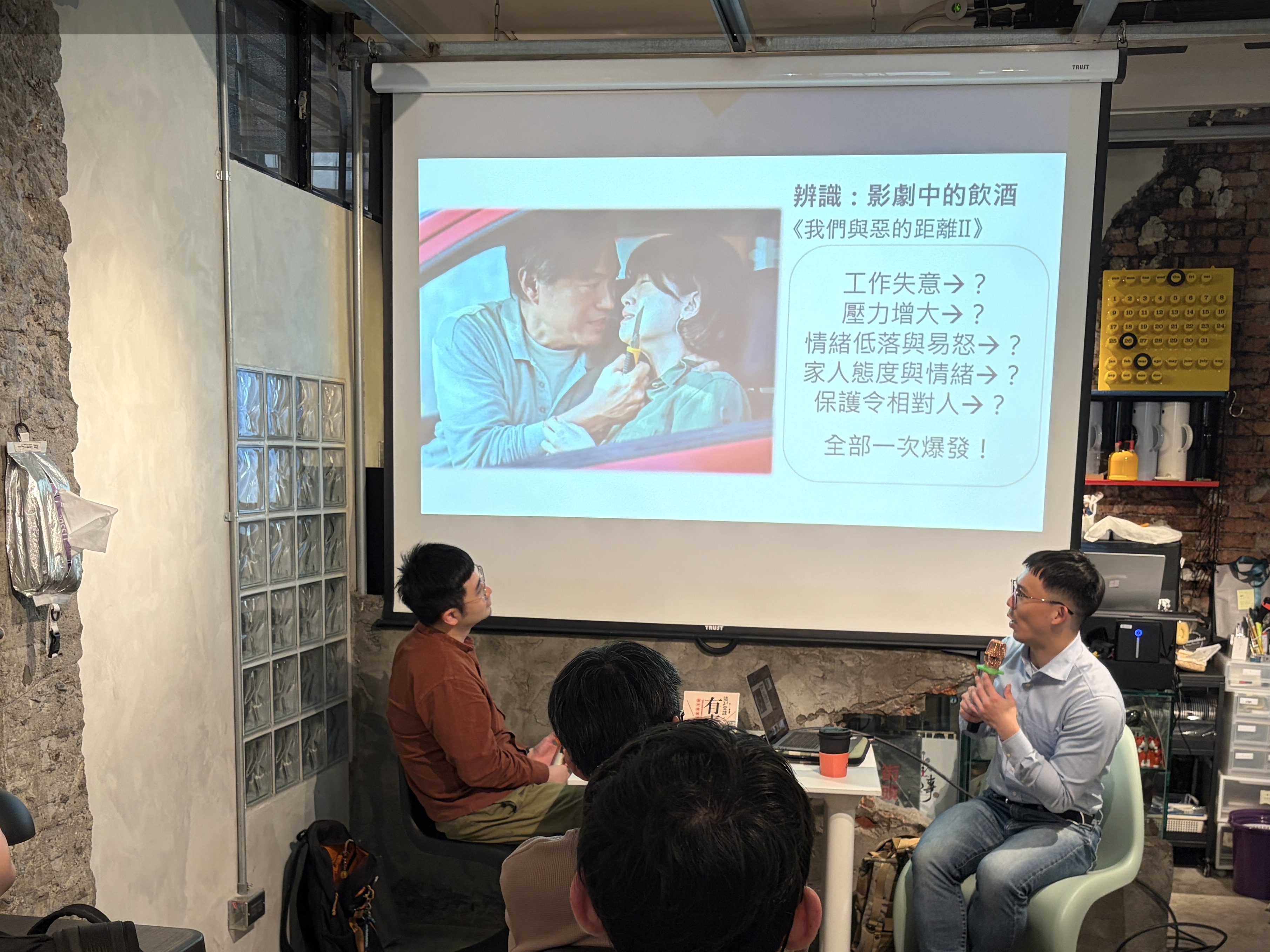 A discussion event in a cozy venue featuring a presentation on a screen. Two speakers are engaged in conversation, while an audience listens attentively. The background displays decorative elements and shared content on the walls.