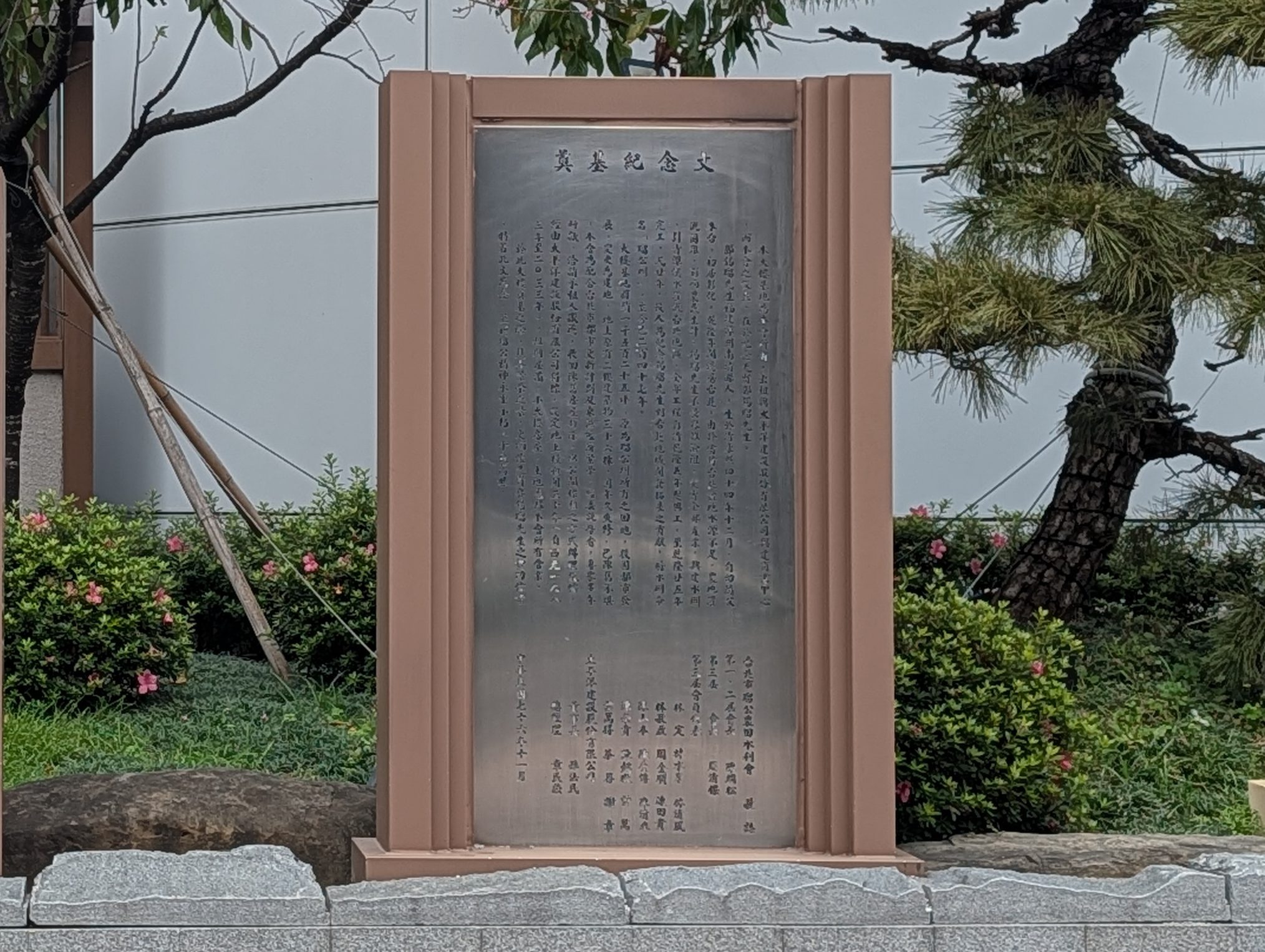 一個開放空間標示牌,旁邊有一塊金屬紀念碑,背景有花朵和松樹,設置於城市街道旁