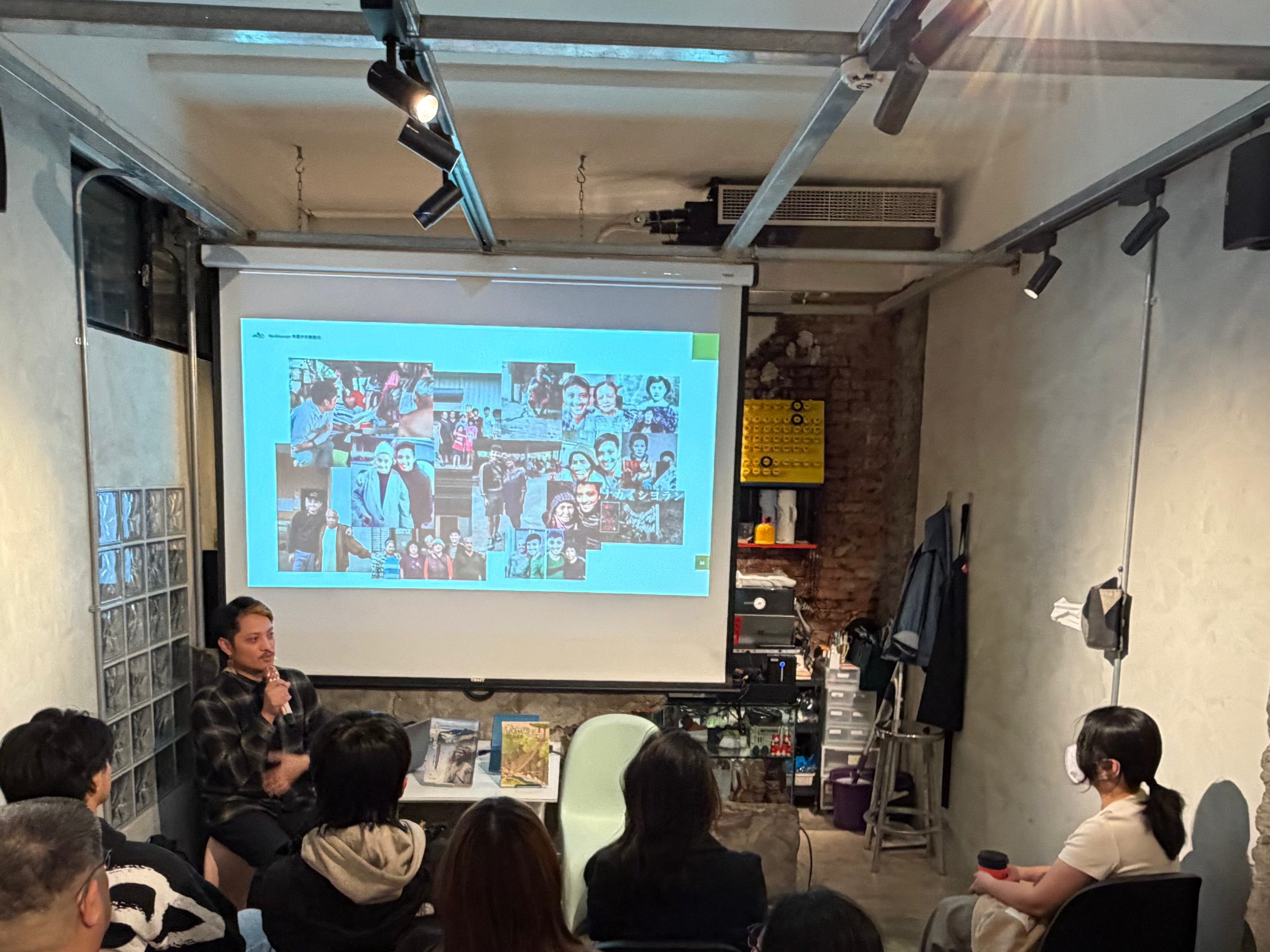 一群人坐在室內的講座中，前方有投影螢幕正在播放圖片，周圍環繞著書架和裝飾物。